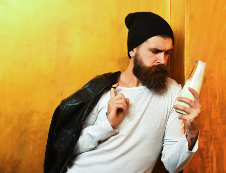 Bearded man, long beard. Brutal caucasian serious unshaven hipster holding glass jar of milk with black leather jacket on shoulder, hat and white shirt on golden studio background, copy spaceの写真素材