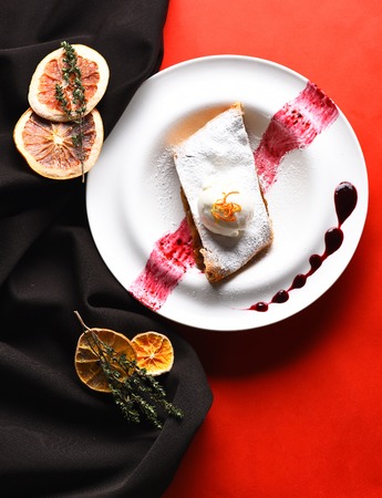 Delicious apple strudel with vanilla ice cream and berry sauce on white plate with dried orange fruit slices and herbs on black fabric on red backgroundの写真素材