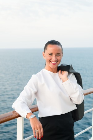 pretty business woman or happy cute girl with smiling face in white blouse, grey jacket, has red manicure on ship deck viewing sea, ocean water on cloudy sky background, traveling and tourismの写真素材