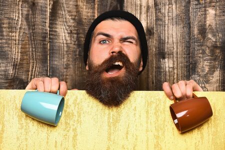 Bearded man, long beard. Brutal caucasian funny unshaven hipster in black hat with yellow paper sheet holding colorful cups on brown vintage wooden backgroundの写真素材