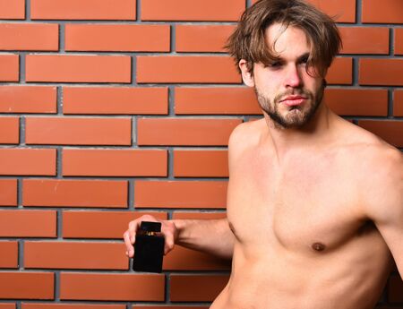 Bearded man, short beard. Caucasian sexy macho man with moustache show six packs and abs on muscle torso on athletic body holding perfume on brown brick wall studio background, copy spaceの写真素材