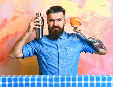 Bearded man, long beard. Brutal caucasian tattooed serious hipster with moustache in shirt doing tropical alcohol fresh cocktail with shaker on blue checkered plaid on colorful texture backgroundの写真素材
