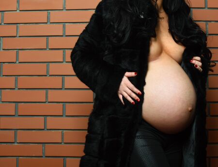female belly of pregnant naked woman in black fur on brown brick wall studio backgroundの写真素材
