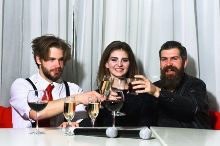 Friends celebrating at karaoke party on white curtain. Two bearded men, hipsters with beards, holding microphones for pretty girl or beautiful woman for singingの写真素材