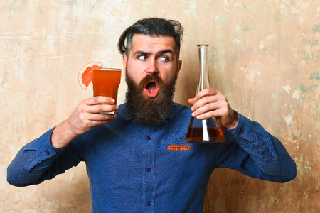 Bearded man, long beard. Brutal caucasian surprised hipster with moustache in denim shirt holding tropical alcoholic fresh cocktail with orange piece and glass tube or flask on texture backgroundの写真素材