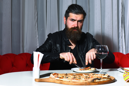 Bearded man or caucasian hipster with long beard and moustache in black leather jacket eating pizza with knife and fork served with glass of red wine in pizzeria, cafe or restaurantの写真素材