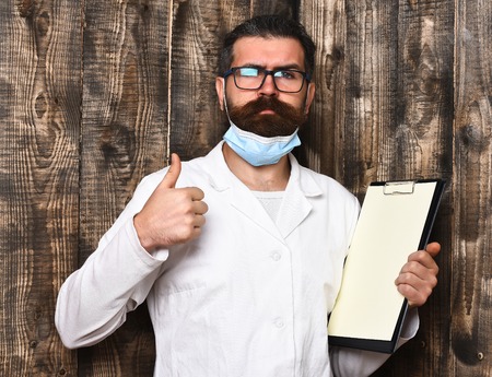 Bearded man, long beard. Brutal caucasian doctor or unshaven hipster, postgraduate student holding clipboard in medical gown and mask on brown wooden vintage studio background. Medicine conceptの写真素材