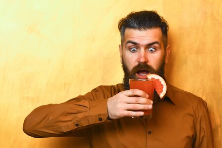 Bearded man, long beard. Brutal caucasian surprised hipster with moustache in brown shirt holding tropical alcoholic fresh cocktail with orange piece on golden texture backgroundの写真素材