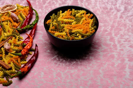 Colorful dried fusilli, italian pasta in bowl plate, with chilly pepper, garlic, cinnamon sticks for cooking vegetarian food on pink background, copy spaceの写真素材