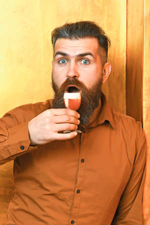 Bearded man, long beard. Brutal caucasian surprised hipster with moustache in brown shirt holding alcoholic red shot on colorful texture backgroundの写真素材