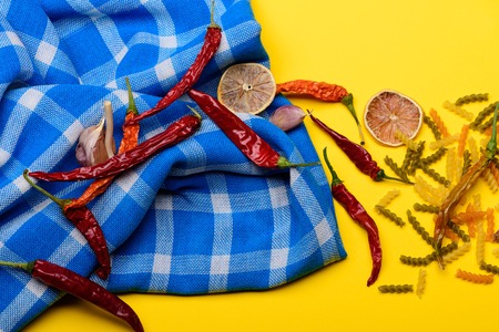 Dried red chilly peppers, orange slices, garlic on blue plaid tablecloth and fusilli pasta, vegetarian food and spices for cooking vegetarian food on yellow backgroundの写真素材