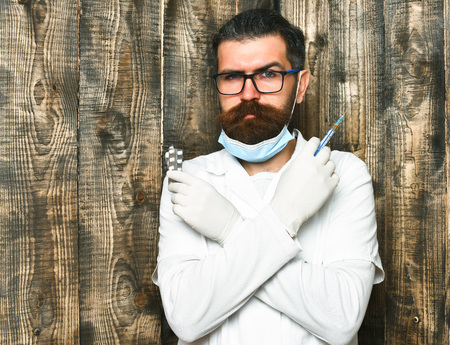 Bearded man, long beard. Brutal caucasian doctor or unshaven hipster, postgraduate student in medical gown, gloves holding syringe and pills on brown vintage wooden studio background. Medicine conceptの写真素材