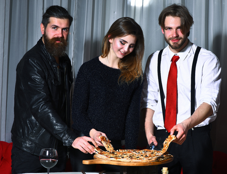 Happy friends, pretty girl and two bearded men, hipsters with beards, smiling and eating tasty pizza served on wooden tray at party in pizzeria, cafe or restaurant on white curtainの写真素材