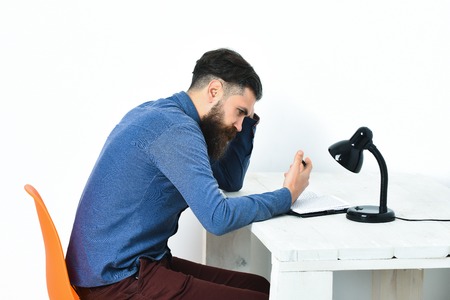 Bearded man, long beard. Brutal caucasian unshaven serious hipster with moustache holding black pen at white vintage wooden table with open notepad and black lamp on it, isolated on white backgroundの写真素材