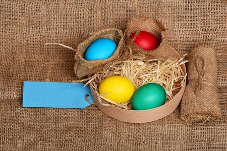 easter colorful eggs painted in bright colors in burlap sack and wooden box with straw and blue shopping tag on sackcloth background, spring holiday celebration, copy spaceの写真素材