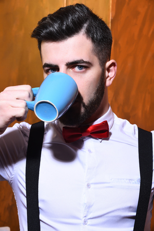 bearded man, long beard, brutal caucasian hipster with moustache in white shirt and suspenders with bow-tie drinking from blue cup of tea or coffee with serious face on brown studio wall backgroundの写真素材