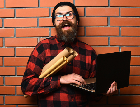 Bearded man, long beard. Brutal caucasian smiling happy unshaven hipster holding laptop and craft paper rolls in red black checkered shirt with hat and glasses on brown brick wall studio backgroundの写真素材