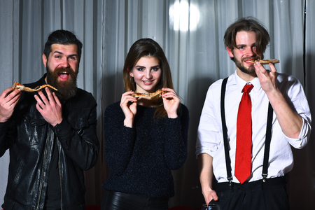 Happy friends, pretty girl and two bearded men, hipsters with beards, smiling and eating tasty pizza slices at party in pizzeria, cafe or restaurant on white curtainの写真素材