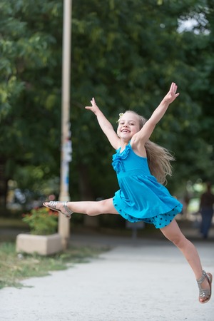 small baby girl or cute happy child with adorable smiling face and bow in blonde hair in blue dress jumping in summer outdoor on blurred backgroundの写真素材