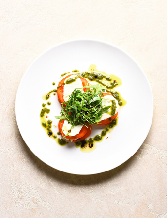 Tasty tomatoes sliced with mozzarella cheese and fresh green arugula salad on plate with spices, herbs on white backgroundの写真素材