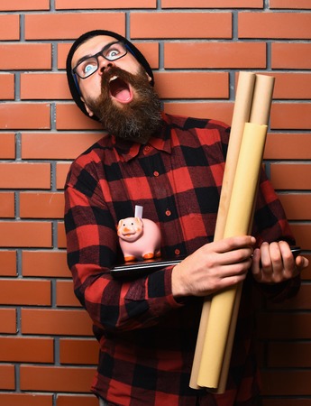 Bearded man, long beard. Brutal caucasian shouting unshaven hipster holding craft paper rolls, piggy bank on laptop in checkered shirt with hat and glasses on brown brick wall studio backgroundの写真素材