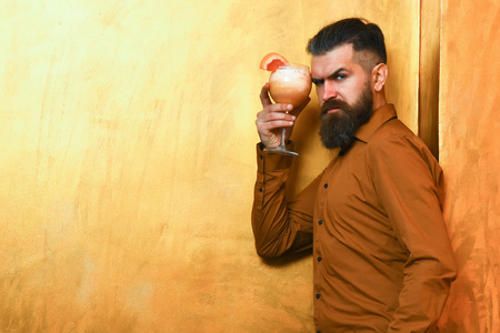 Bearded man, long beard. Brutal caucasian serious hipster with moustache in brown shirt holding tropical alcoholic fresh cocktail with orange piece on golden texture background, copy spaceの写真素材