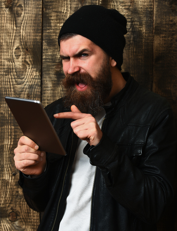 Bearded man, long beard. Brutal caucasian funny unshaven hipster holding tablet in black leather jacket on brown vintage wooden studio backgroundの写真素材