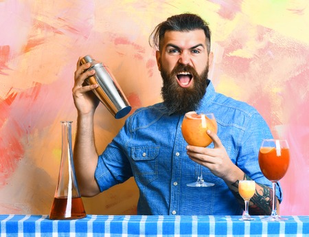 Bearded man, long beard. Brutal caucasian hipster with moustache in denim shirt doing cocktails in shaker and alcohol shot with bar stuff on blue checkered plaid on colorful texture backgroundの写真素材