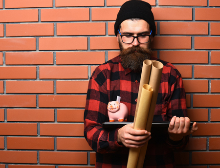 Bearded man, long beard. Brutal caucasian serious unshaven hipster holding craft paper rolls, piggy bank on laptop in checkered shirt with hat and glasses on brown brick wall studio backgroundの写真素材