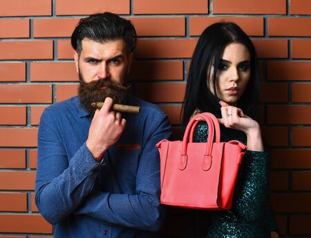 serious couple of bearded man, long beard, brutal caucasian hipster with moustache and pretty woman or sexy girl with brunette hair, makeup in green sequins dress hold bag and cigar on brick wallの写真素材