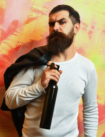 Bearded man, long beard. Brutal caucasian serious unshaven hipster holding bottle and smoking cigar with black leather jacket on shoulder and white shirt on colorful studio backgroundの写真素材