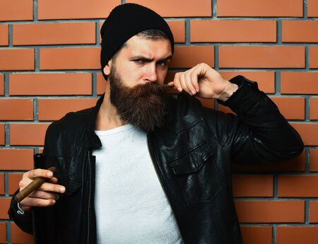 Bearded man, long beard. Brutal caucasian serious unshaven hipster smoking cigar with black leather jacket, hat and white shirt on brown brick wall studio backgroundの写真素材