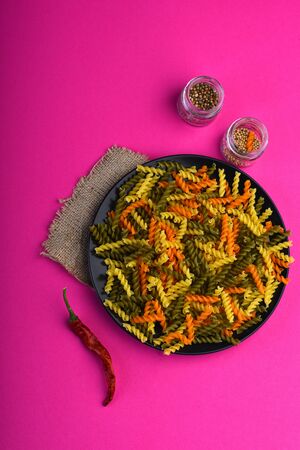 Colorful dried fusilli pasta on black plate on sackcloth with red chilly pepper and spices in glass jars on pink background. Ingredients for cooking italian or vegetarian foodの写真素材