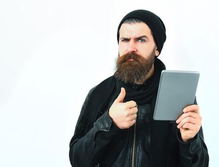 Bearded man, long beard. Brutal caucasian serious unshaven hipster holding tablet in black leather jacket, hat and scarf isolated on white studio backgroundの写真素材