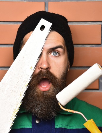 bearded worker man, long beard, brutal caucasian hipster with moustache holding various building tools: saw, hammer, roller paint with surprised face on brick wall backgroundの写真素材