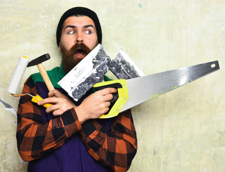 bearded man, long beard, brutal caucasian hipster with moustache holding various building tools: saw, hammer, roller paint with surprised face on studio wall backgroundの写真素材