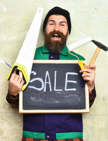 bearded foreman, long beard, brutal caucasian hipster with moustache holding various building tools: saw, hammer, roller paint, board with inscription sale with happy face on studio wall backgroundの写真素材