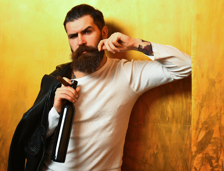 Bearded man, long beard. Brutal caucasian serious unshaven hipster holding bottle and smoking cigar with black leather jacket on shoulder and white shirt on golden studio backgroundの写真素材