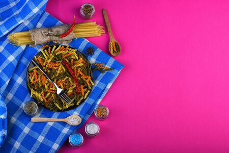 Colorful dried fusilli and spaghetti pasta, red chilly peppers on black plate with metal fork and wooden spoons on blue plaid tablecloth on pink background, copy spaceの写真素材