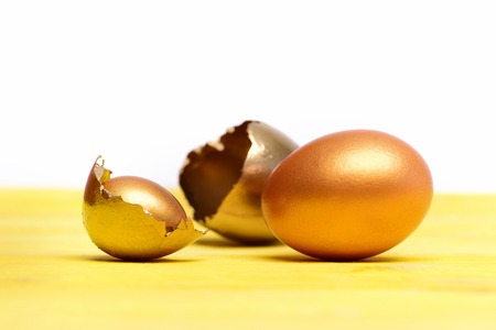 retirement, business and finance, golden broken egg with shell, eggshell, easter food painted in gold metallic color on yellow wood isolated on white background, future life, luxury and successの写真素材
