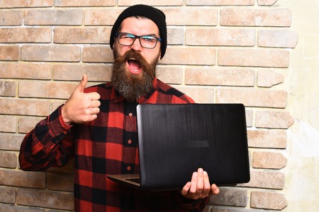 Bearded man, long beard. Brutal caucasian happy unshaven hipster holding laptop in red black checkered shirt with hat and glasses on beige brick wall studio backgroundの写真素材