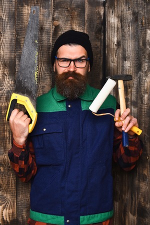 bearded painter, long beard, brutal caucasian hipster with moustache holding various building tools: saw, hammer, roller paint with serious face on brown wooden studio backgroundの写真素材