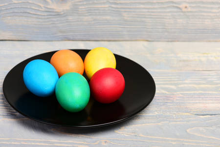 colorful painted handmade eggs in black round kitchen plate on grey wooden background, happy easter...
