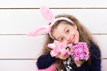 greeting and celebration, cute girl in bunny ears, happy small child with curly hair, tulip flower, pink bouquet on white wooden background, womens or mothers day spring holidayの写真素材