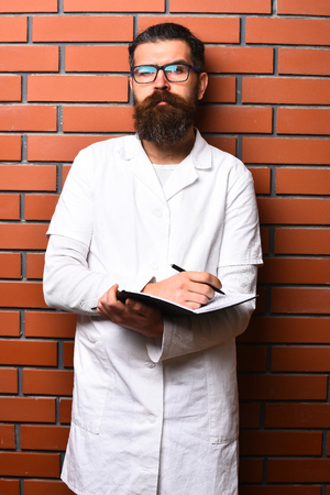 Bearded man, long beard. Brutal caucasian doctor or unshaven hipster, postgraduate student holding notebook with pen in medical gown and glasses on brown brick wall studio background. Medicine conceptの写真素材