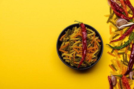 Colorful fusilli, dried italian pasta, with red chilly peppers in black bowl, ingredients for cooking vegetarian food on yellow background, copy spaceの写真素材