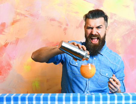 Bearded man, long beard. Brutal caucasian happy tattooed hipster with moustache in denim shirt doing tropical alcohol fresh cocktail with shaker on blue checkered plaid on colorful texture backgroundの写真素材