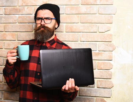 Bearded man, long beard. Brutal caucasian serious unshaven hipster holding laptop with mag or cup in red black checkered shirt with hat and glasses on beige brick wall studio backgroundの写真素材