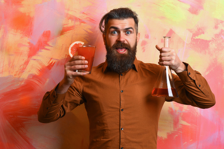 Bearded man, long beard. Brutal caucasian happy hipster with moustache in brown shirt holding tropical alcoholic fresh cocktail with orange piece and glass tube or flask on colorful texture backgroundの写真素材
