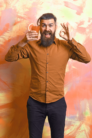 Bearded man, long beard. Brutal caucasian smiling happy hipster with moustache in brown shirt holding tropical alcoholic fresh cocktail with orange piece on colorful texture backgroundの写真素材